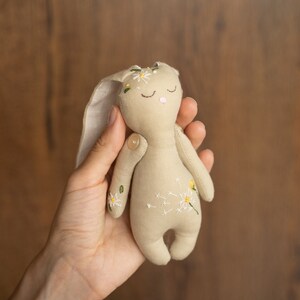 May include: A beige fabric bunny toy with embroidered white flowers and green leaves. The bunny has closed eyes and a pink nose.