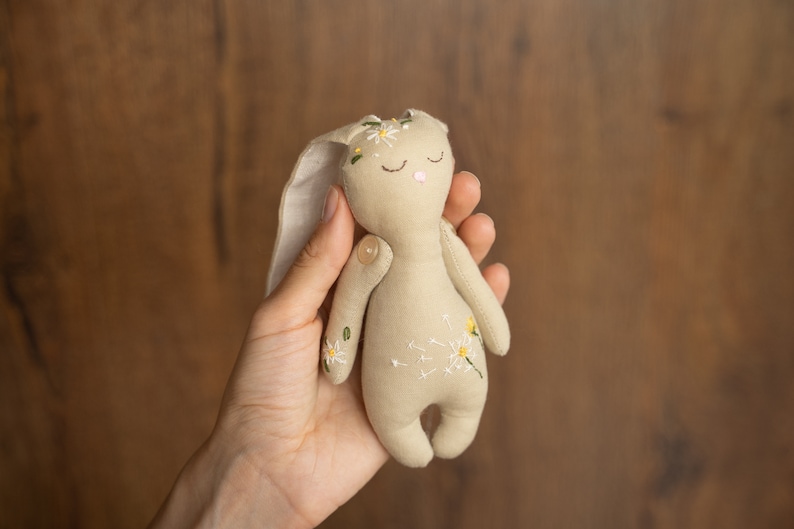 May include: A beige fabric bunny toy with embroidered white flowers and green leaves. The bunny has closed eyes and a pink nose.