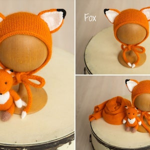 May include: A knitted orange fox-themed bonnet with white and black ear details and matching chin ties, accompanied by a small stuffed orange fox toy with white chest fur and brown paws, and a long orange knitted wrap or scarf accessory.