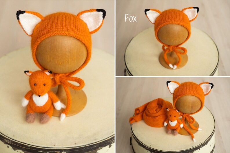 May include: A knitted orange fox-themed bonnet with white and black ear details and matching chin ties, accompanied by a small stuffed orange fox toy with white chest fur and brown paws, and a long orange knitted wrap or scarf accessory.