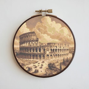 May include: A brown wooden embroidery hoop with a beige and brown cross-stitch design of the Colosseum in Rome. The design includes a cloudy sky and a few small figures in the foreground.