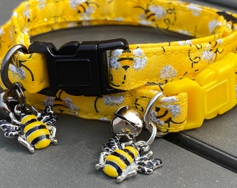 May include: Yellow cat collar with a bee pattern and two bee charms. The collar has a black buckle and a yellow buckle. The charms are silver with black stripes.
