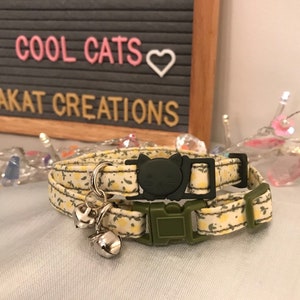 May include: A cat collar with a yellow floral pattern, a black cat-shaped buckle, and a silver bell. The collar has a green buckle and a matching green cat-shaped buckle. The background includes the text "COOL CATS" and "#MIAKAT CREATIONS".