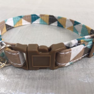 May include: A cat collar with a geometric pattern in teal, brown, white, and yellow. The collar has a brown plastic buckle and a small silver bell. The collar is approximately 0.5 inches wide.