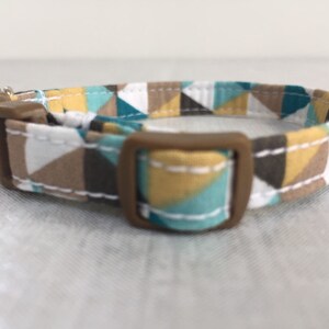 May include: A geometric patterned cat collar featuring a brown plastic buckle and a small silver bell. The fabric design includes triangles in teal, brown, white, and yellow. The collar is designed for a cat.