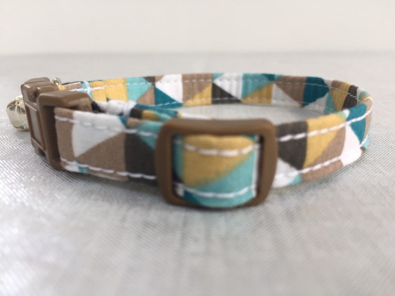 May include: A geometric patterned cat collar featuring a brown plastic buckle and a small silver bell. The fabric design includes triangles in teal, brown, white, and yellow. The collar is designed for a cat.