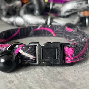 Może przedstawiać: Czarny i różowy obroża dla kota z wzorem pajęczyny i dzwoneczkiem. Obroża ma plastikową klamrę.