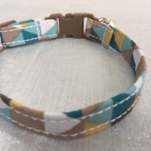 May include: A patterned pet collar featuring a geometric design in teal, white, brown, and yellow. The collar has a brown plastic buckle and a metal loop for attaching a leash. The fabric is stitched with white thread.