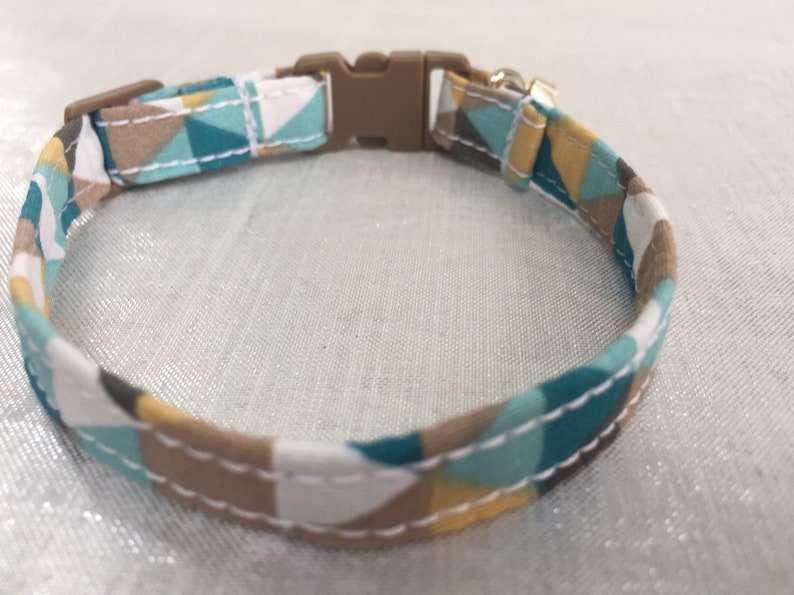 May include: A patterned pet collar featuring a geometric design in teal, white, brown, and yellow. The collar has a brown plastic buckle and a metal loop for attaching a leash. The fabric is stitched with white thread.