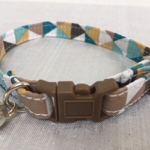 May include: A cat collar with a geometric pattern in teal, brown, white, and yellow. The collar has a brown plastic buckle and a silver bell attached. The collar is approximately 0.5 inches wide.