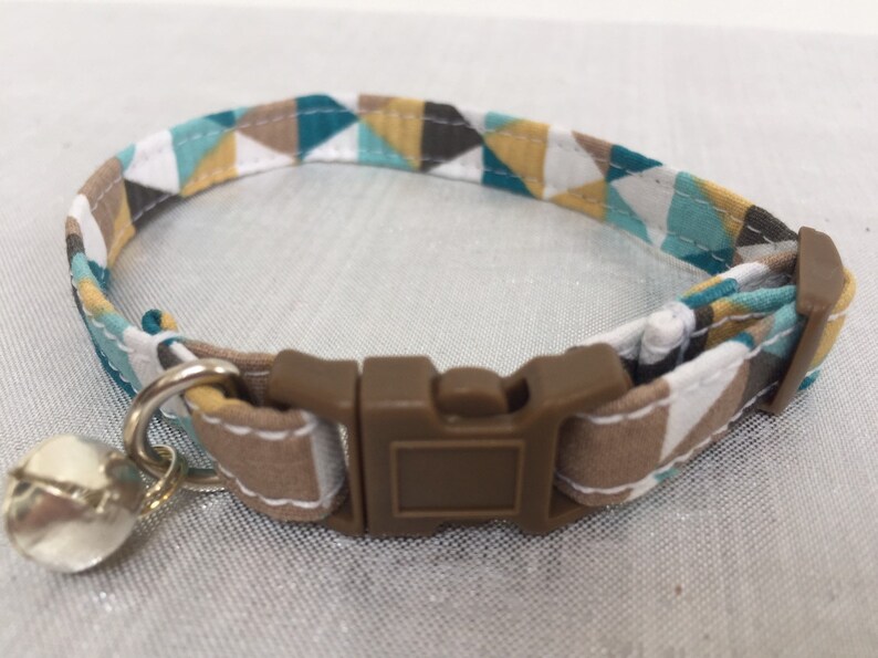 May include: A cat collar with a geometric pattern in teal, brown, white, and yellow. The collar has a brown plastic buckle and a silver bell attached. The collar is approximately 0.5 inches wide.