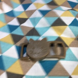 May include: A brown cat-shaped buckle, featuring a cat face design. The buckle is made of plastic and has a rectangular shape with two slots. The background is a fabric with a geometric pattern of triangles in blue, white, brown, and yellow.