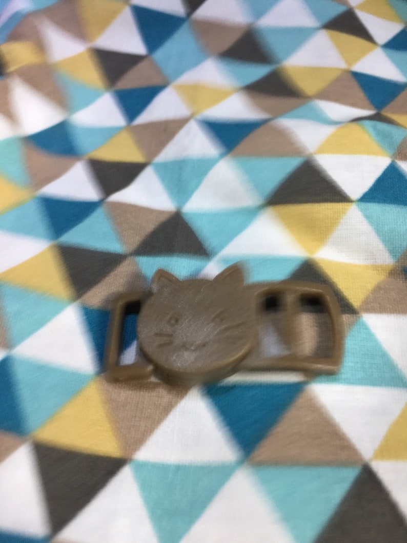 May include: A brown cat-shaped buckle, featuring a cat face design. The buckle is made of plastic and has a rectangular shape with two slots. The background is a fabric with a geometric pattern of triangles in blue, white, brown, and yellow.