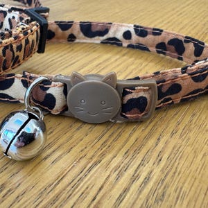 Könnte beinhalten: Ein Katzenhalsband mit Leopardenmuster in Braun- und Schwarztönen. Das Halsband hat eine braune Kunststoffschnalle mit einem Katzen-Gesicht-Design und eine silberne Glocke. Das Halsband liegt auf einer Holzoberfläche.