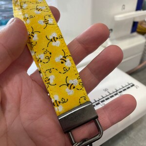 Op de afbeelding: Een gele stoffen sleutelhanger met een bijenpatroon en een zilveren metalen sleutelring.