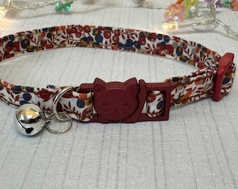 Puede incluir: Un collar para gatos con estampado floral, hebilla roja con forma de gato y cascabel plateado. El collar presenta un patrón de flores rojas, azules y blancas sobre fondo crema. La hebilla y el cierre son rojos.