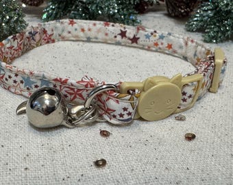 Puede incluir: Un collar para gatos estampado con una hebilla color crema y una campana plateada. El collar presenta una tela blanca con estrellas rojas, azules y amarillas. La hebilla tiene forma de cara de gato.