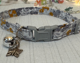 Puede incluir: Un collar para gato con estampado floral en gris y blanco, con hebilla de plástico gris. Tiene una campana plateada y un dije de mariposa. El patrón incluye elementos florales amarillos y grises.