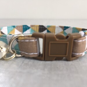 May include: A cat collar with a geometric pattern in teal, brown, white, and yellow. The collar has a brown plastic buckle and a silver bell. The collar is made of fabric and is designed for a cat.
