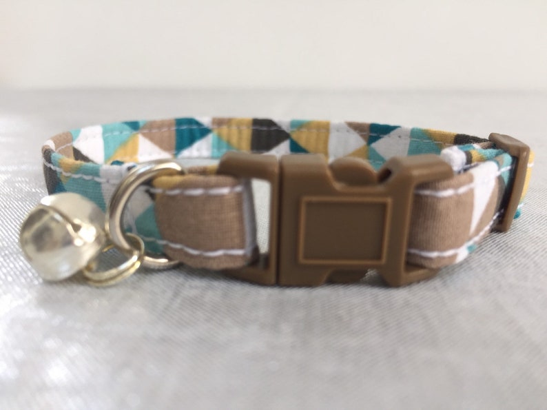 May include: A cat collar with a geometric pattern in teal, brown, white, and yellow. The collar has a brown plastic buckle and a silver bell. The collar is made of fabric and is designed for a cat.
