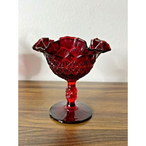 Puede incluir: Un cuenco de pedestal de vidrio rojo rubí con un borde con volantes. El cuenco tiene un patrón de diamantes y se asienta sobre una base redonda. El tallo tiene un diseño decorativo.