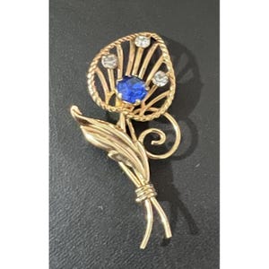 Puede incluir: Broche floral dorado con una piedra preciosa azul en el centro y tres piedras claras más pequeñas. El broche tiene un diseño detallado de hojas y tallos, con un borde retorcido en forma de cuerda alrededor de la flor. Estilo vintage.