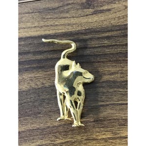 Könnte beinhalten: Eine goldfarbene Metallbrosche in Form einer stilisierten Katze. Die Katze steht mit über den Rücken gebogenem Schwanz. Die Brosche hat eine glänzende Oberfläche und ist auf einem Holzgrund platziert.