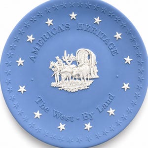May include: A round light blue decorative plate featuring white relief details. The center depicts a covered wagon pulled by oxen with a cactus. Text reads AMERICA'S HERITAGE and The West - By Land. The border is decorated with white stars.