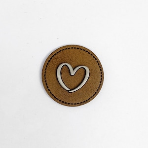 May include: A brown leather patch with a silver heart-shaped design. The heart is outlined in silver and has a slightly curved shape.