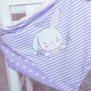 Op de afbeelding: Een paars-wit gestreept babydekentje met een wit konijnontwerp en witte sterren in de onderste hoek. Het dekentje is gevouwen en over een witte stoel gelegd.