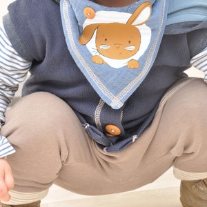 Op de afbeelding: Een baby draagt een blauwe en bruine vest met capuchon met een blauwe en witte slabbetje met een bruine konijn illustratie. Het slabbetje heeft een witte streep en een bruine knoop. De baby draagt een bruine broek en bruine schoenen.