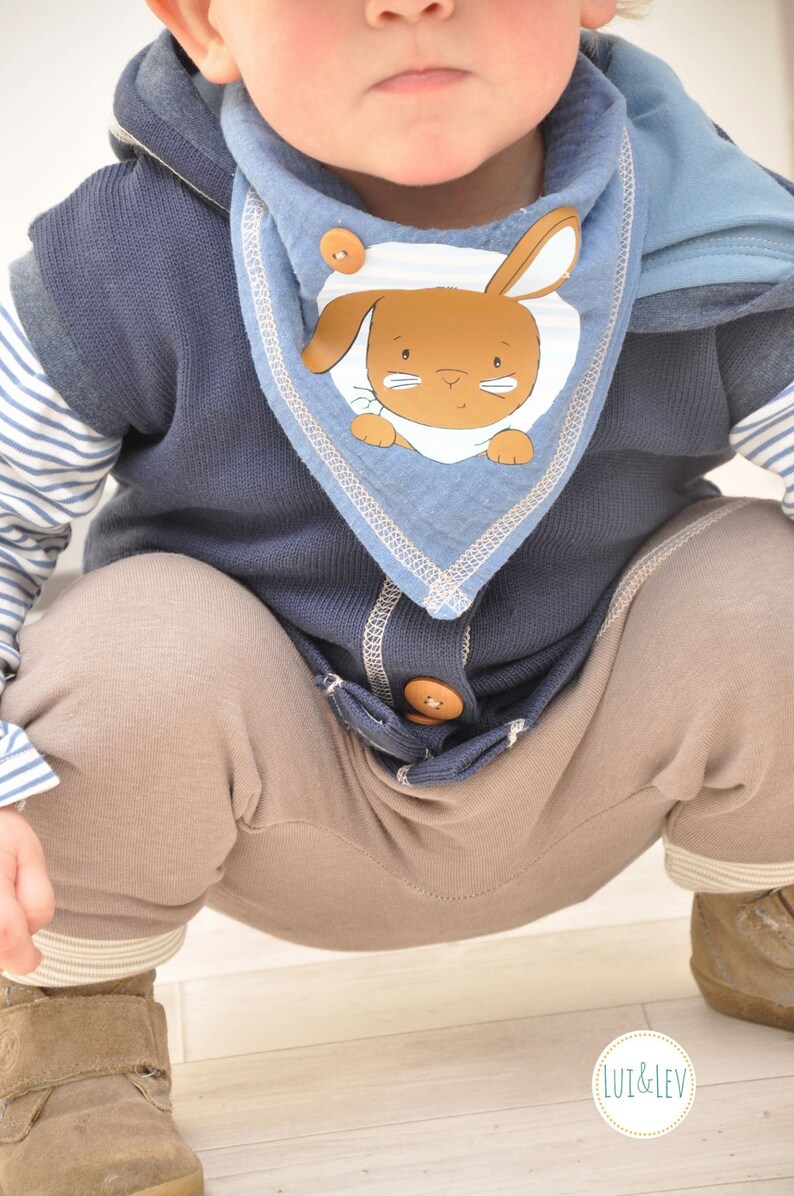 Op de afbeelding: Een baby draagt een blauwe en bruine vest met capuchon met een blauwe en witte slabbetje met een bruine konijn illustratie. Het slabbetje heeft een witte streep en een bruine knoop. De baby draagt een bruine broek en bruine schoenen.