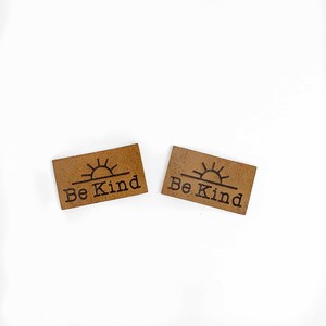 May include: Two brown leather patches with a sun graphic and the text "Be Kind" in black.