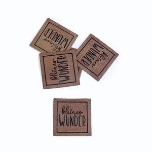 May include: Four brown leather labels with the text "kleines WUNDER" in black lettering. The labels are scattered on a white background.