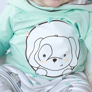 May include: A baby wearing a mint green hooded sweatshirt with a white puppy graphic and matching striped pants with a puppy print. The sweatshirt has a star-shaped button.