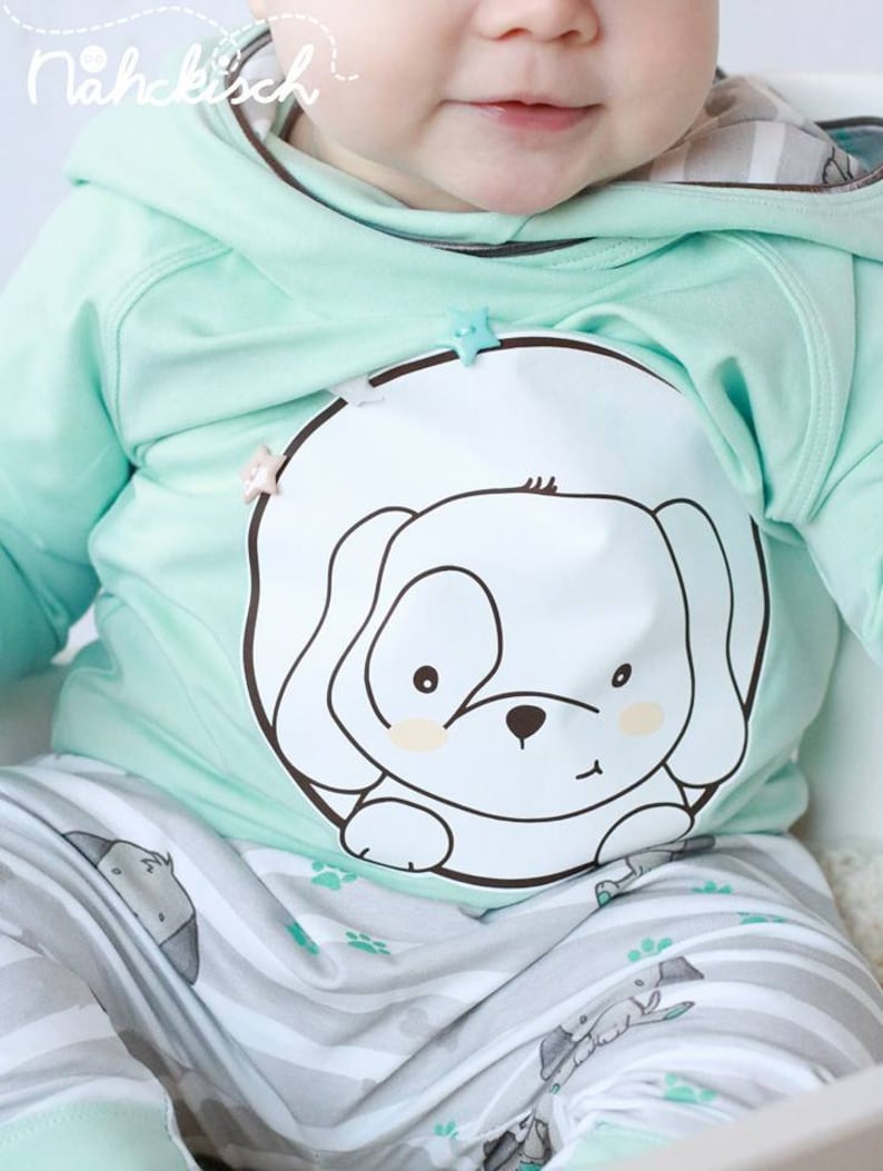 May include: A baby wearing a mint green hooded sweatshirt with a white puppy graphic and matching striped pants with a puppy print. The sweatshirt has a star-shaped button.