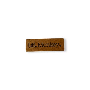 Könnte beinhalten: Ein braunes Leder-Etikett mit dem Text "Little Monkey" in schwarzen Buchstaben und einem kleinen Herzsymbol.
