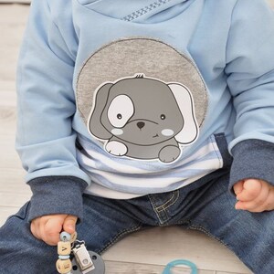 May include: A baby wearing a light blue hooded sweatshirt with a gray and white puppy applique. The baby is holding a wooden bead pacifier clip with the name "Novan" spelled out.