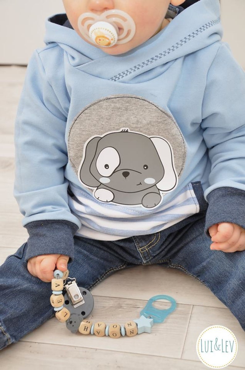May include: A baby wearing a light blue hooded sweatshirt with a gray and white puppy applique. The baby is holding a wooden bead pacifier clip with the name "Novan" spelled out.