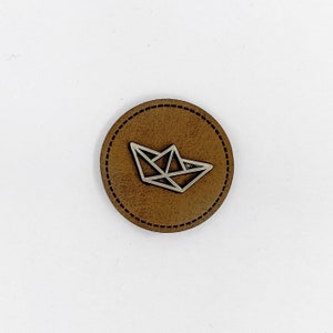 May include: A circular, brown leather patch with a stitched edge. A silver geometric paper boat design is in the centre. The patch is set against a white background.