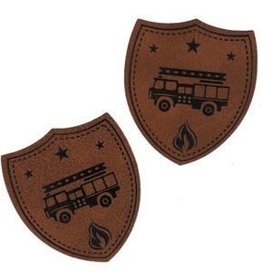 Può includere: Due toppe in pelle marrone a forma di scudo con cuciture nere sui bordi. Ogni toppa presenta una silhouette nera di un camion dei pompieri con una scala e una fiamma. Ci sono tre stelle nere su ogni toppa.