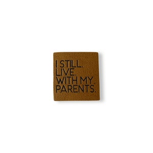 Könnte beinhalten: Ein quadratischer, brauner Lederaufnäher mit dem geprägten Satz "I STILL LIVE. WITH MY PARENTS." in Schwarz. Der Aufnäher hat eine strukturierte Oberfläche und ist auf weißem Hintergrund isoliert.