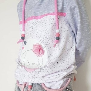 Op de afbeelding: Een grijs en wit hoodie met een roze konijn afbeelding en roze en witte stippen. De hoodie heeft roze trekkoordjes met witte kralen. Het model draagt ook grijze shorts met roze bies en witte kralen op het trekkoord.