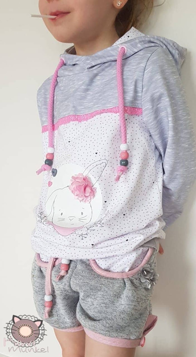 Op de afbeelding: Een grijs en wit hoodie met een roze konijn afbeelding en roze en witte stippen. De hoodie heeft roze trekkoordjes met witte kralen. Het model draagt ook grijze shorts met roze bies en witte kralen op het trekkoord.
