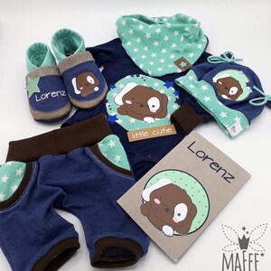 May include: A baby gift set with a blue and brown bodysuit, pants, hat, booties, and a bib. The set features a brown puppy design with white stars and the text "little cutie". The bib and hat are personalized with the name "Lorenz".