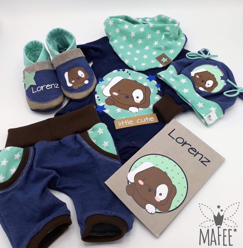 May include: A baby gift set with a blue and brown bodysuit, pants, hat, booties, and a bib. The set features a brown puppy design with white stars and the text "little cutie". The bib and hat are personalized with the name "Lorenz".