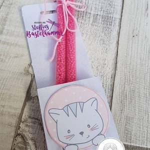 Pode incluir: Um conjunto de presentes com dois tubos transparentes cheios de contas rosa, amarrados com barbante rosa. O conjunto inclui um cart&atilde;o com uma ilustra&ccedil;&atilde;o de gato de desenho animado e o texto "Steffies Bastelhammer".