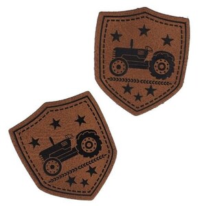 Op de afbeelding: Twee bruine leren patches in de vorm van schilden met zwarte stiksels rond de randen. Elke patch heeft een zwart silhouet van een tractor met vijf sterren en een rij tarwehalmen.