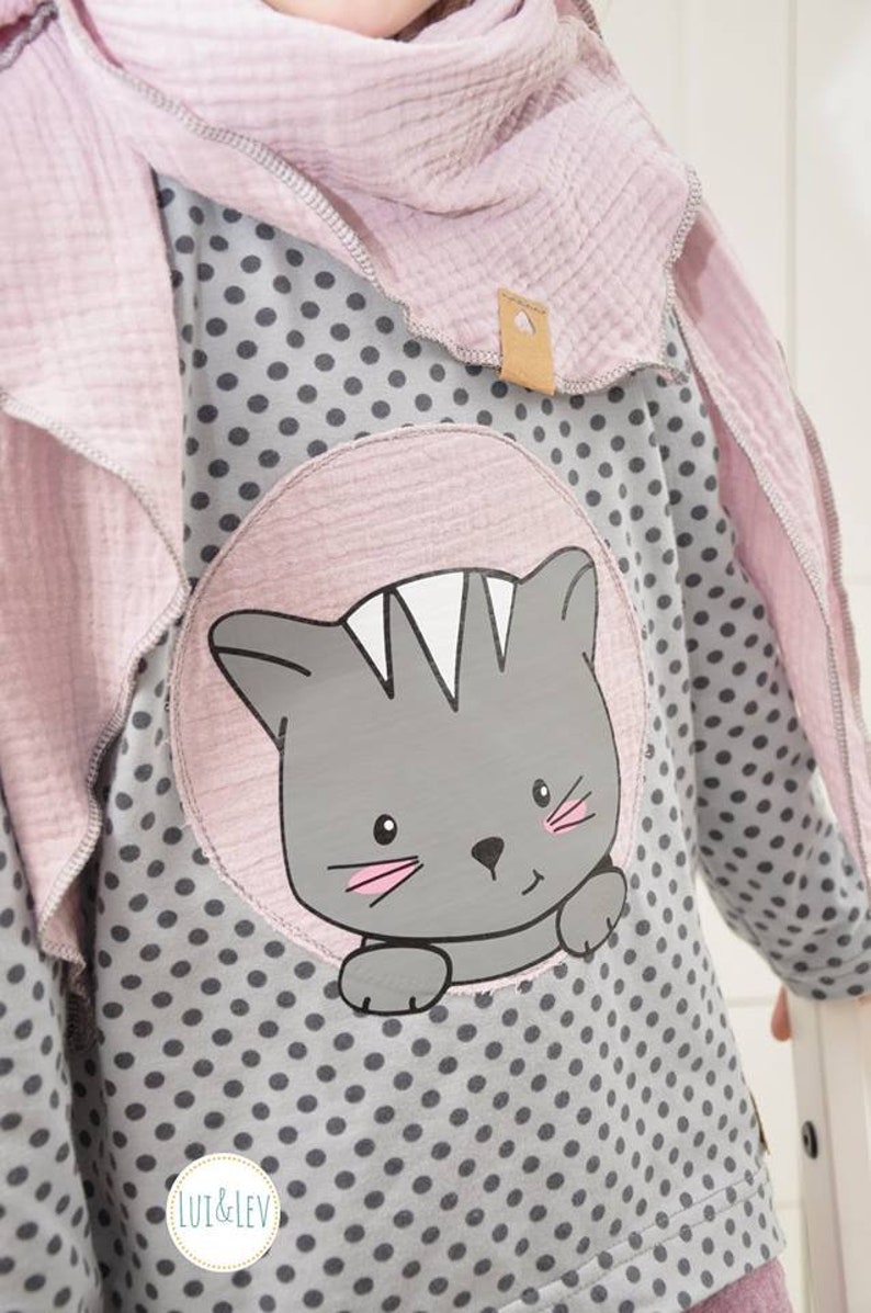 Pode incluir: Uma camisa de manga comprida cinza e branca com bolinhas e um desenho de gato rosa. A camisa tem uma gola rosa e um cachecol rosa a condizer. O texto "LUI & LEV" est&aacute; impresso na camisa.