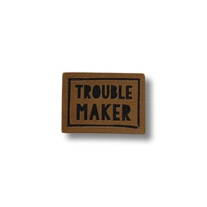 Puede incluir: Un parche de cuero marrón con texto negro que dice "TROUBLE MAKER" en una fuente en negrita y en bloque.